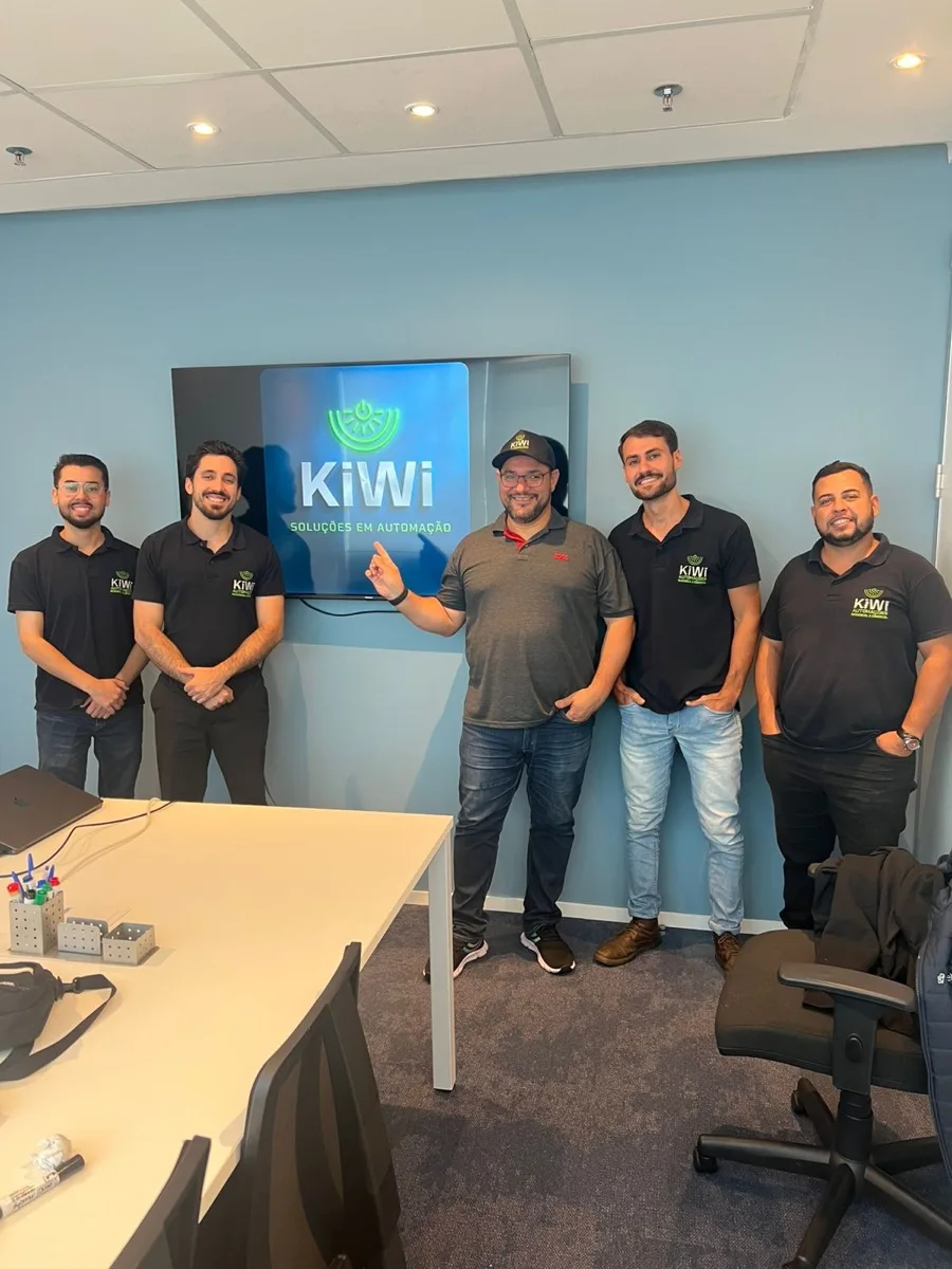 Equipe da KiWi Automação reunida no escritório, em frente ao painel da marca